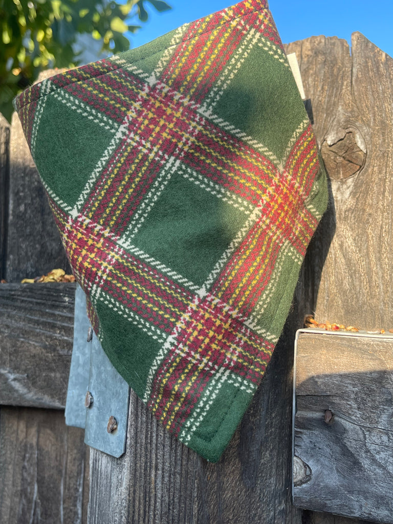 Classic flannel dog bandana resting on a rustic wood fence in warm light. Plaid fabric shows green, cranberry, and gold tones