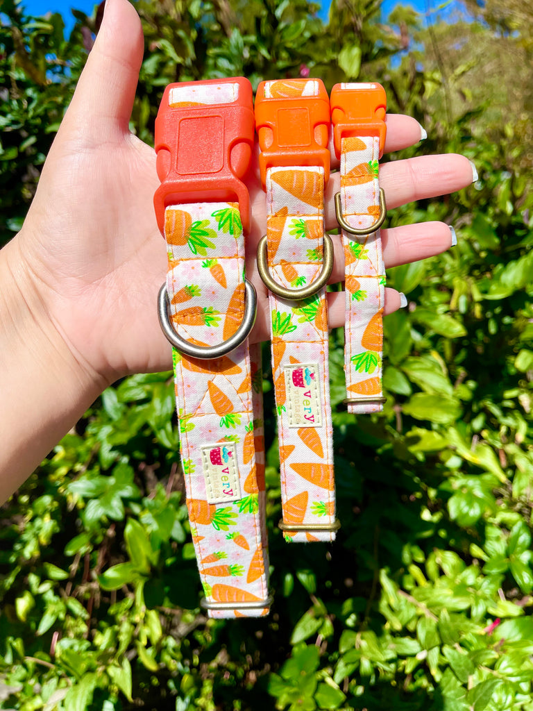 Handmade Carrot Blossom Dog Collar displayed on a hand outdoors against a lush green ivy backdrop. This eco-friendly, organic cotton collar features a playful design with vibrant orange carrots, green stems, and pink flowers on a white background. Adjustable and available in multiple sizes, this collar is perfect for small to large dogs, adding a seasonal touch to your pup’s wardrobe for Easter or spring.