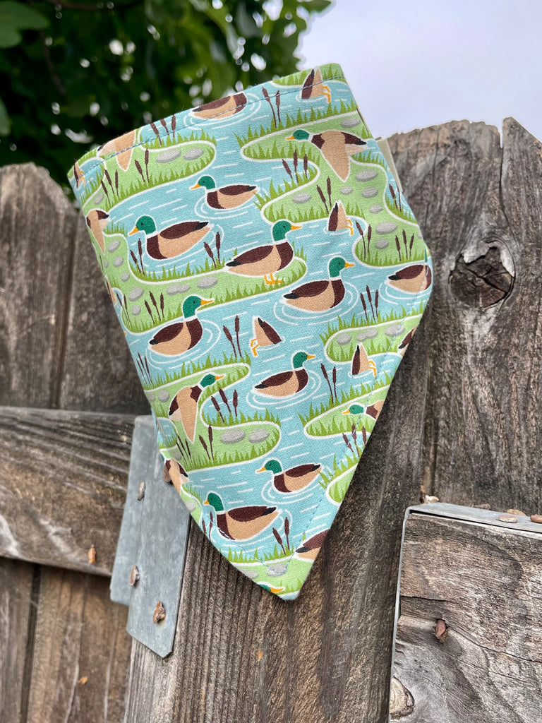 Duck-themed pet bandana with ponds and lily pads, shown hanging on rustic fence