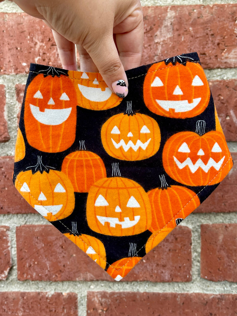 Hand holding a Halloween dog bandana with a pattern of orange jack-o’-lanterns on a black background. Brick wall in the background