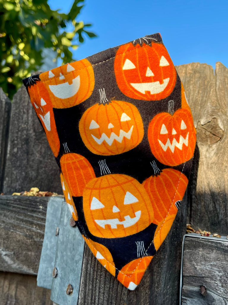 Pumpkin-patterned Halloween bandana displayed on a wooden fence with morning sunlight. Bright orange jack-o’-lanterns pop against the black