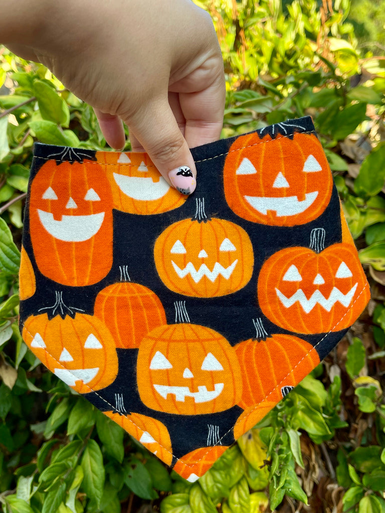 Close-up of the Jack o’ Lantern bandana held in front of leafy greenery. Features smiling pumpkins in different expressions on dark fabric