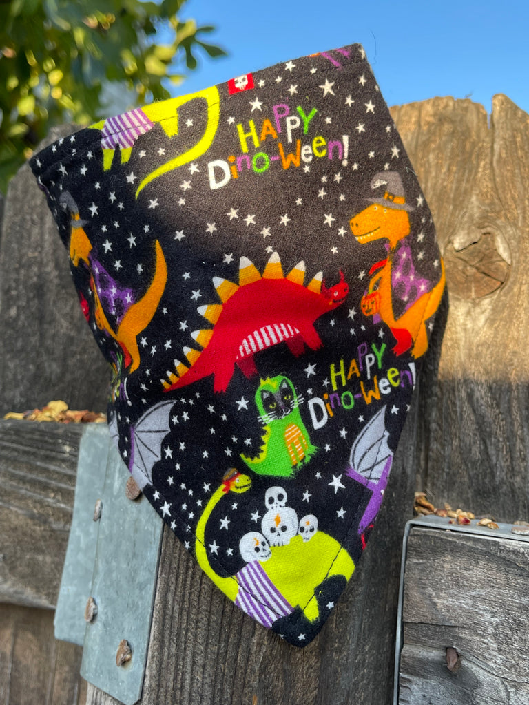 Halloween-themed bandana draped over a rustic fence in warm sunlight. Pattern shows dinos, cats, skulls, and “Happy Dino-Ween” lettering on a starry black background