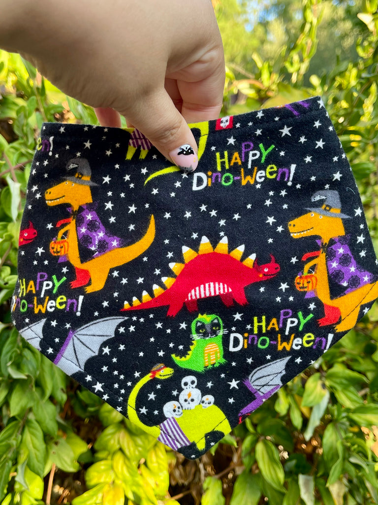 Close-up of the Happy Dinoween bandana held in front of leafy greenery. Shows bright dinos, candy buckets, skeletons, and stars on black fabric