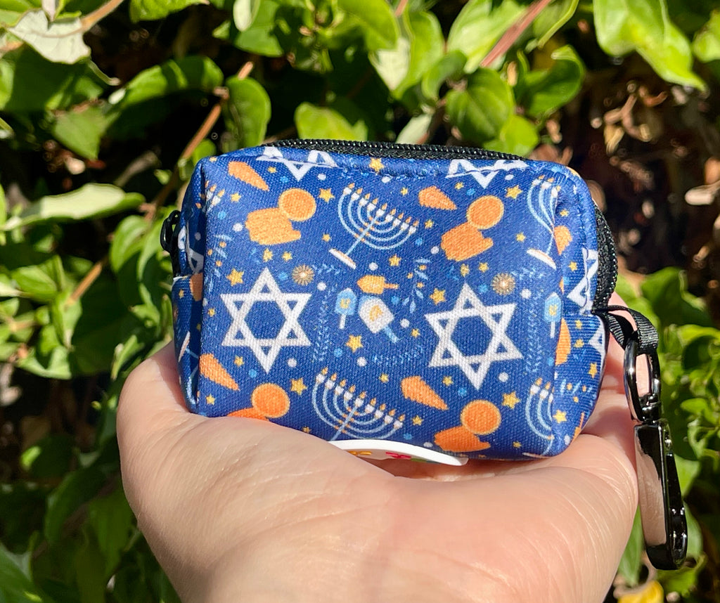 Bright blue poop bag holder with Hanukkah-themed symbols held in front of green leaves; features a festive pattern of dreidels, gelt, and menorahs