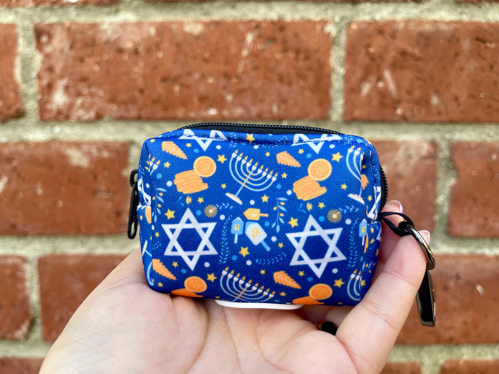 Hand holding a blue Hanukkah-themed dog poop bag holder with menorahs, dreidels, Stars of David, and gold gelt