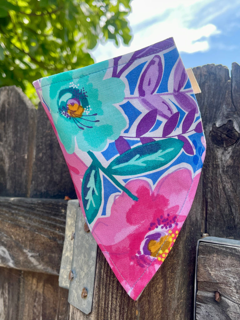 Colorful floral dog bandana with aqua and pink flowers, hanging on wood fence with green leaves and blue sky in background