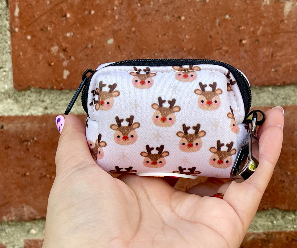 Hand holding a holiday poop bag holder with blushy reindeer faces on a soft white background, shown against a red brick wall.