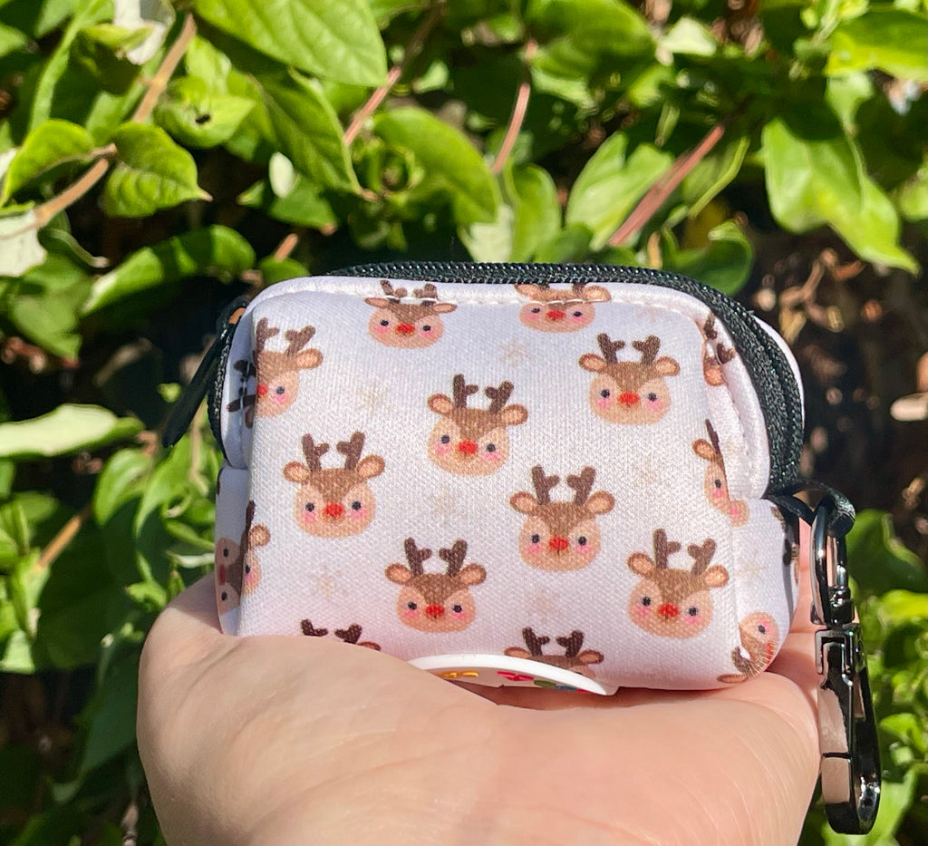 Hand holding the poop bag holder in front of green foliage; pattern shows festive reindeer heads on soft cream fabric with subtle snowflakes