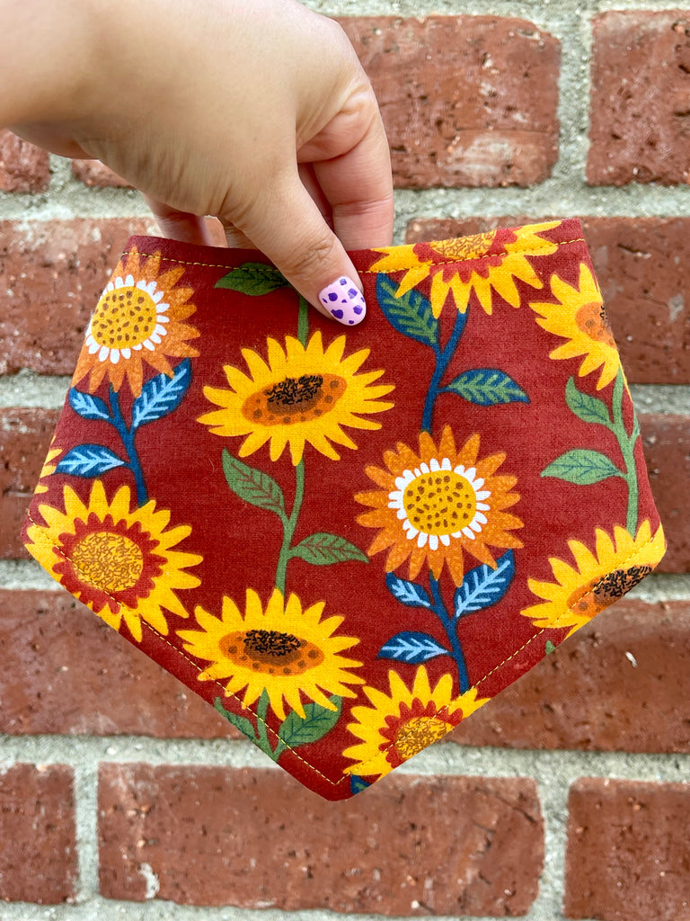 Hand holding a fall dog bandana featuring large yellow sunflowers and green leaves on a rich red background, shown in front of a brick wall