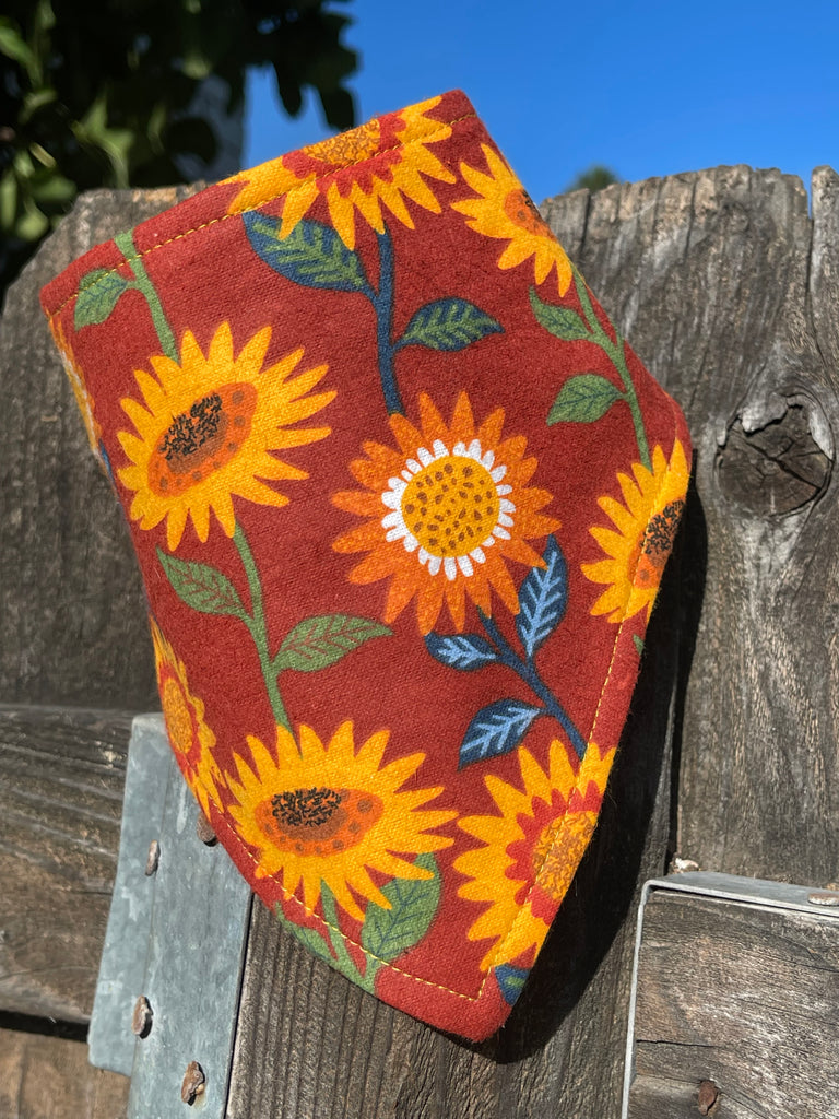 Close-up of sunflower dog bandana hanging on a rustic wood fence; golden floral pattern pops against the red base and bright autumn sun.