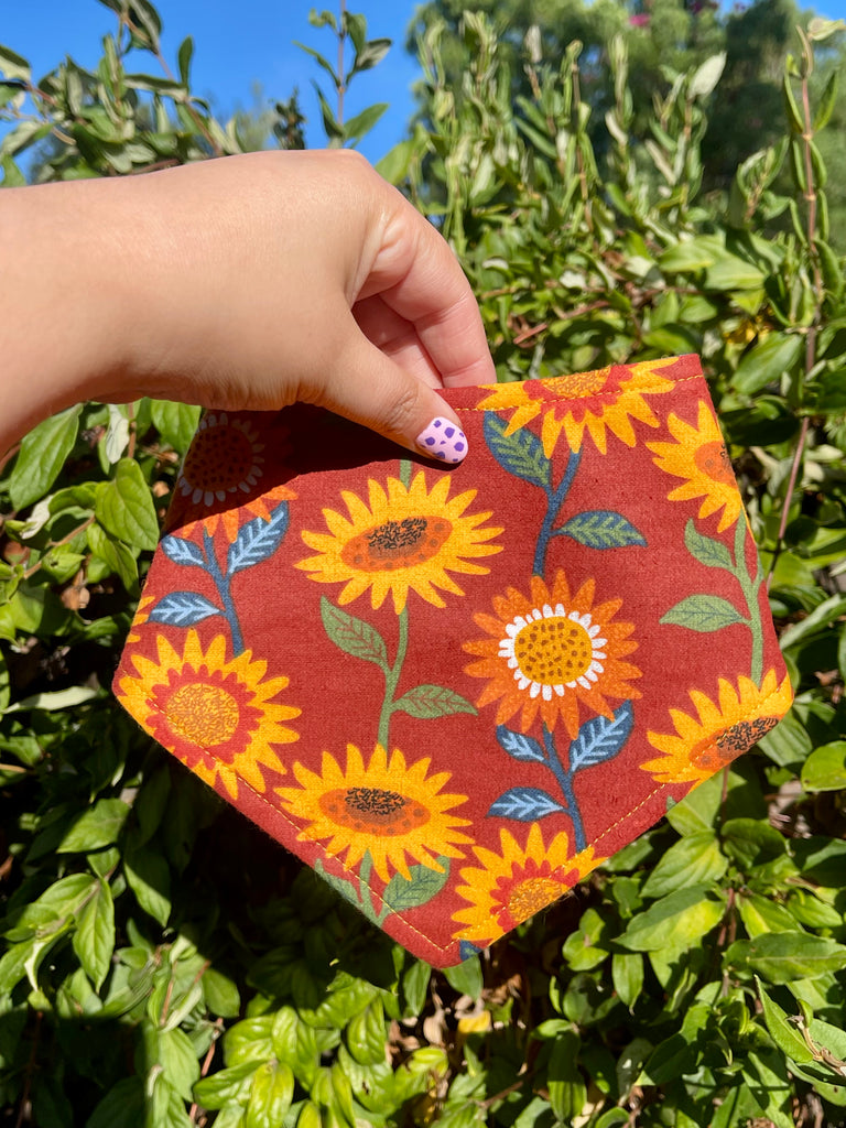 Hand holding sunflower print dog bandana outdoors with vibrant greenery and bright blue sky behind it.