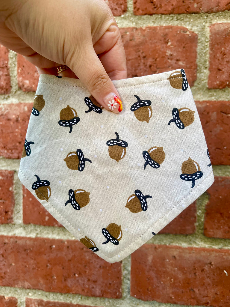 Light tan acorn print bandana held against a red brick wall, highlighting cozy fall colors and curved stitching detail.