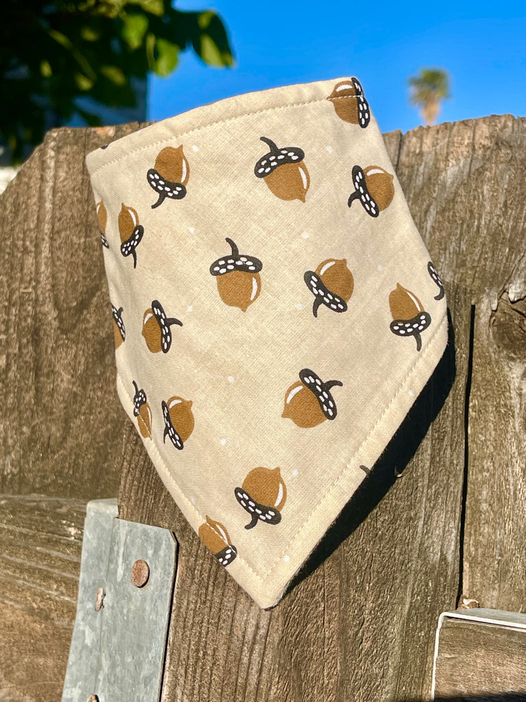 Acorn-themed dog bandana draped on a rustic wooden fence with blue sky above, showing the triangular shape and soft cotton texture.