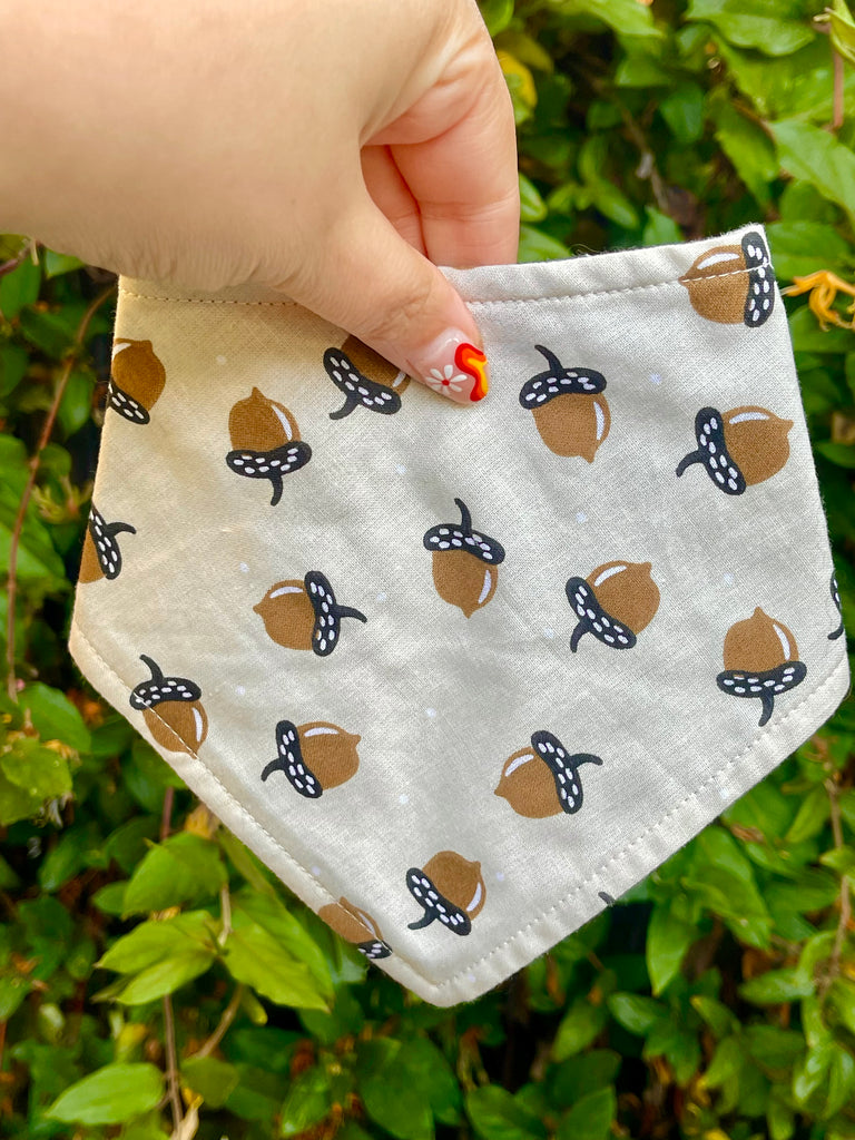 Neutral-toned acorn dog bandana held in front of green leaves, showing autumn-inspired print and handmade stitching.