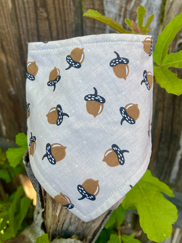Eco-friendly pet bandana with brown acorn pattern, displayed outdoors on a tree stump with leafy foliage for a woodland-inspired fall look.