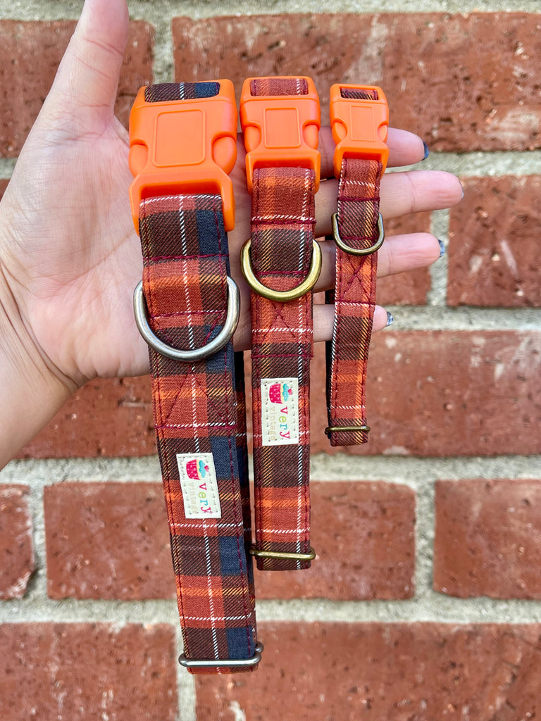 Hand holding orange-buckled fall plaid dog collars against brick wall