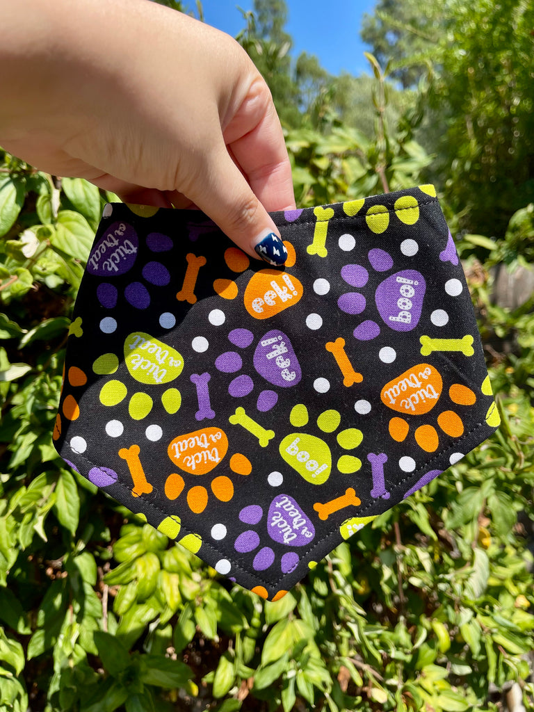 Colorful halloween pet bandana with paw prints and bones that say "trick or treat", "boo", "eek" and held by a hand against a green leafy background.