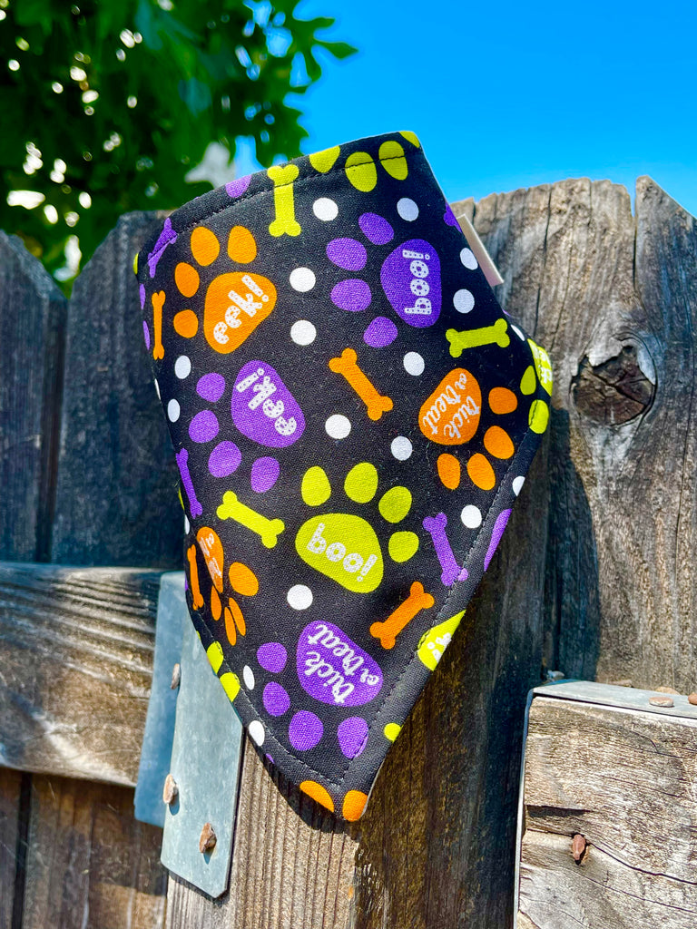 Black halloween dog bandana with orange, purple and lime green paw prints and bones design on a wooden fence
