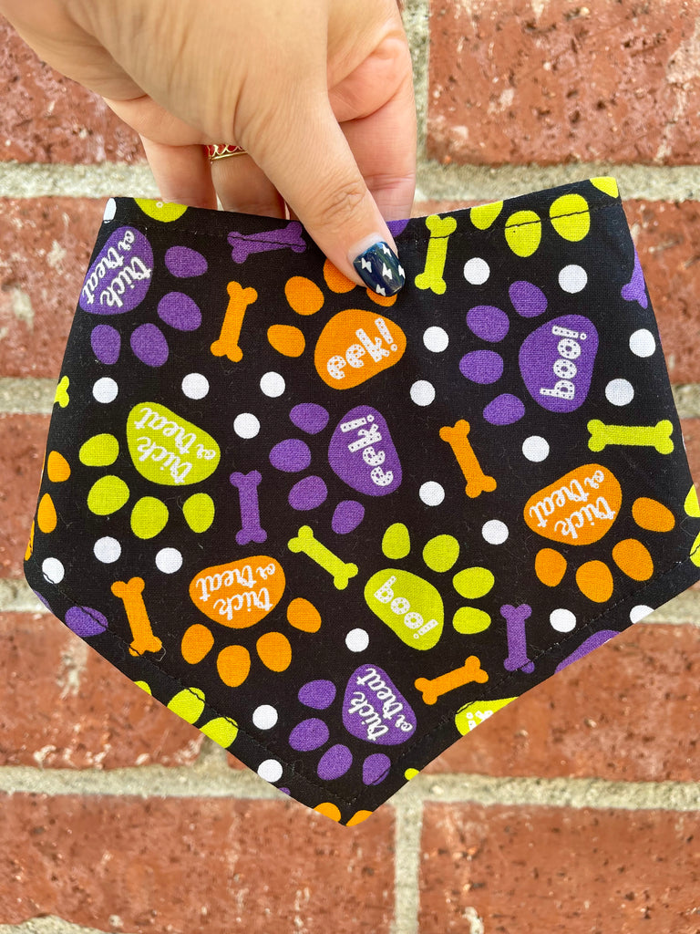Black bandana with colorful paw print and bone pattern for Halloween held by a hand against a brick wall.