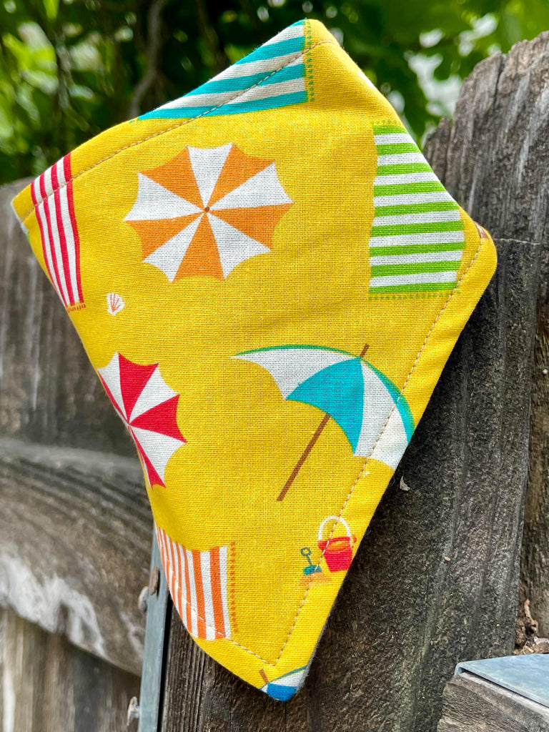 Adjustable beach umbrella dog bandana, folded triangle style over a rustic post, shown in natural light to highlight it's bright summer pattern, artisan-made in LA.