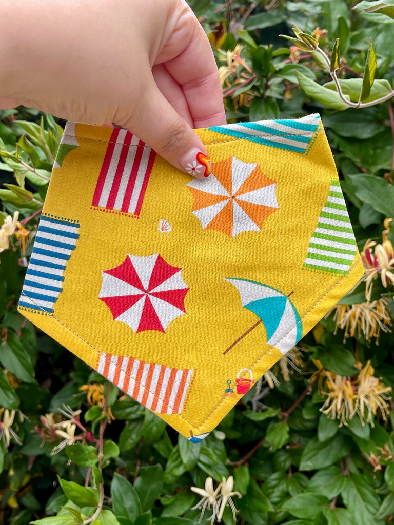 A yellow dog bandana with beach umbrellas and towels, displayed outdoors against a lush green garden background, handmade eco-friendly pet accessory.