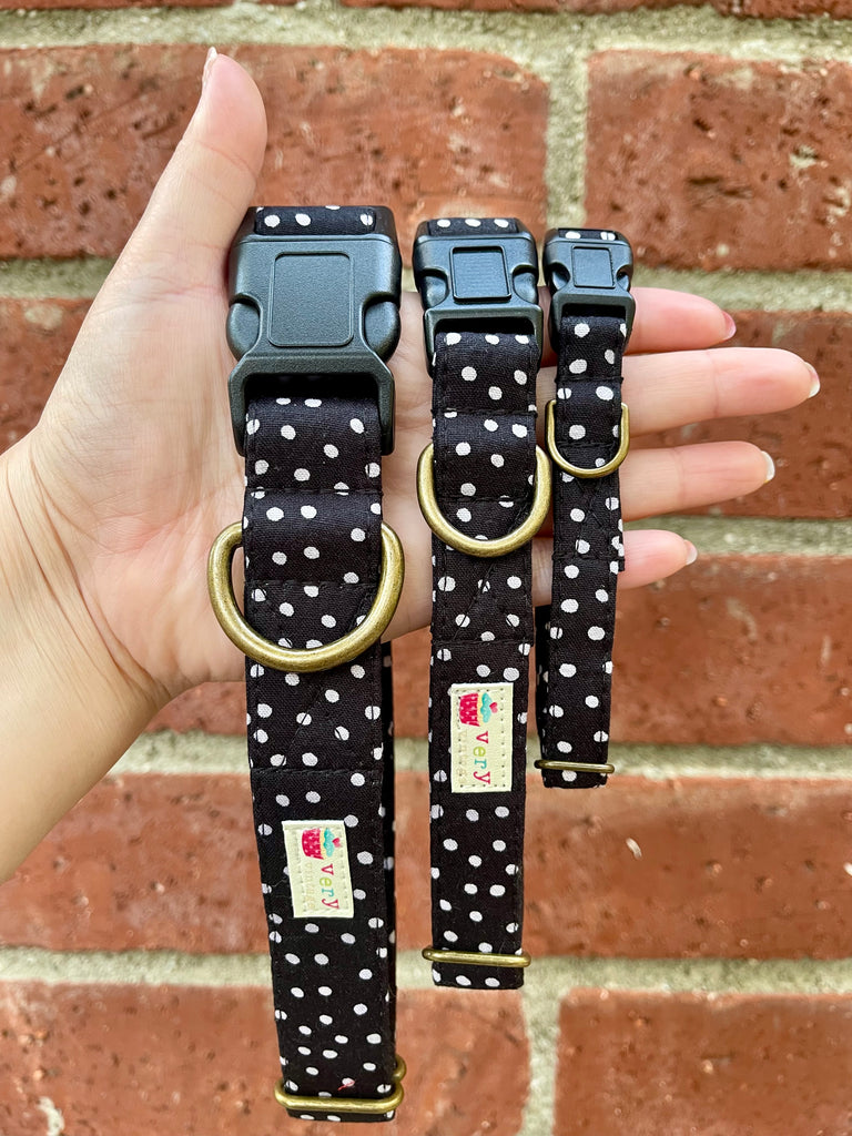 Hand holding a black and white polka dot pet collar in front of a vintage red brick wall; eco-friendly organic cotton with antique brass D-ring and curved buckle.