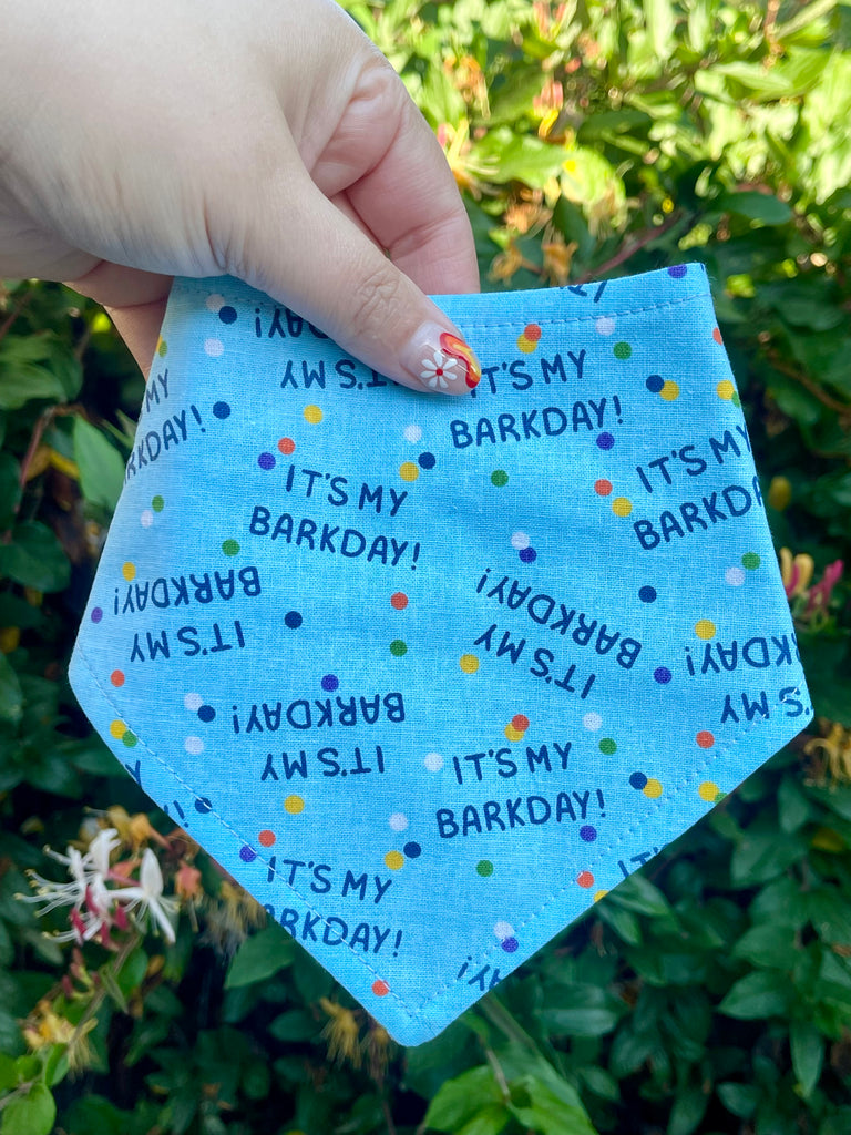 Handmade dog bandana with blue background and rainbow sprinkles, printed with “It’s My Barkday!” phrase. Shown outdoors with leafy green backdrop.