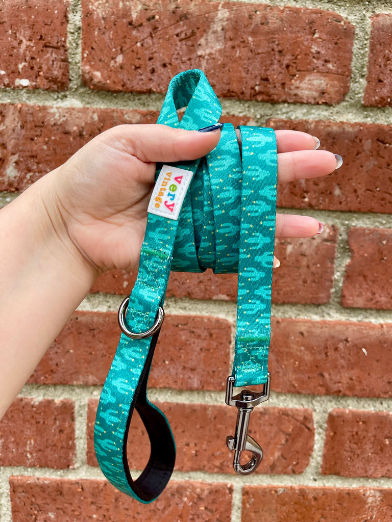 Dog leash with teal cactus print and black hardware held up in front of a red brick wall, showing handle and clip