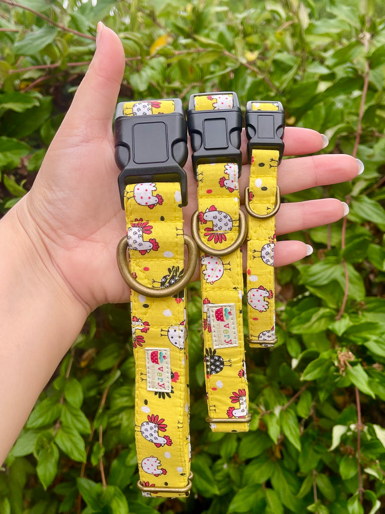 Farm-inspired dog collar with white chickens on yellow fabric and black buckle, held in hand near green garden leaves