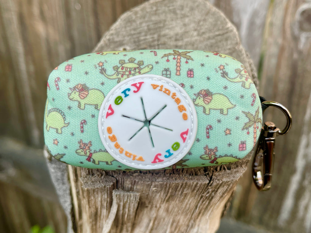 Front view of Christmas Dinos poop bag holder against wood background – shows holiday dino print with “Very Vintage” rubber port and black clip