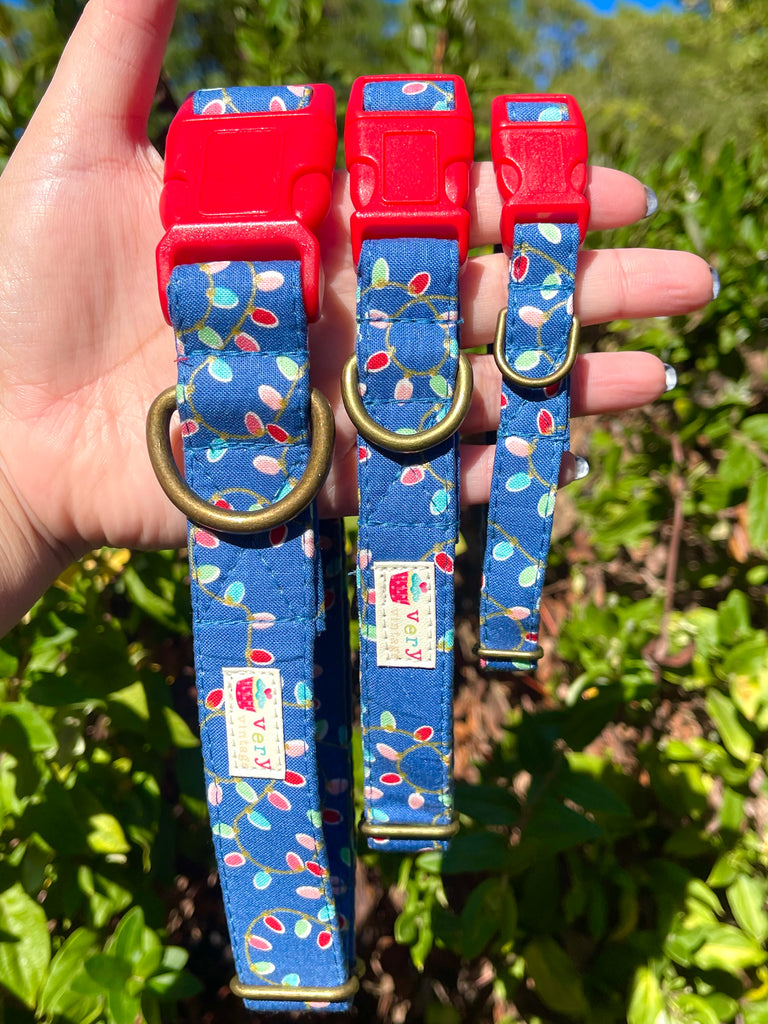 Hand holding three eco-friendly Christmas dog collars in front of greenery – navy blue organic cotton collars with festive light print and brass hardware
