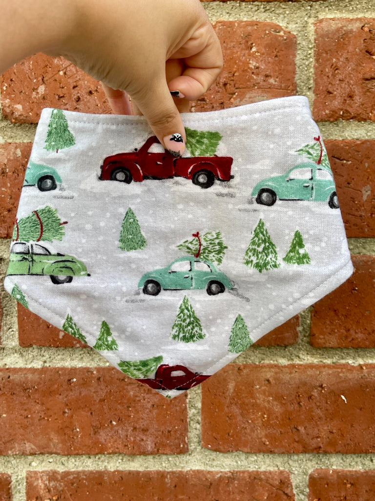 Hand holding a winter dog bandana featuring red trucks and green cars carrying Christmas trees on a snowy gray background. Red brick wall behind.