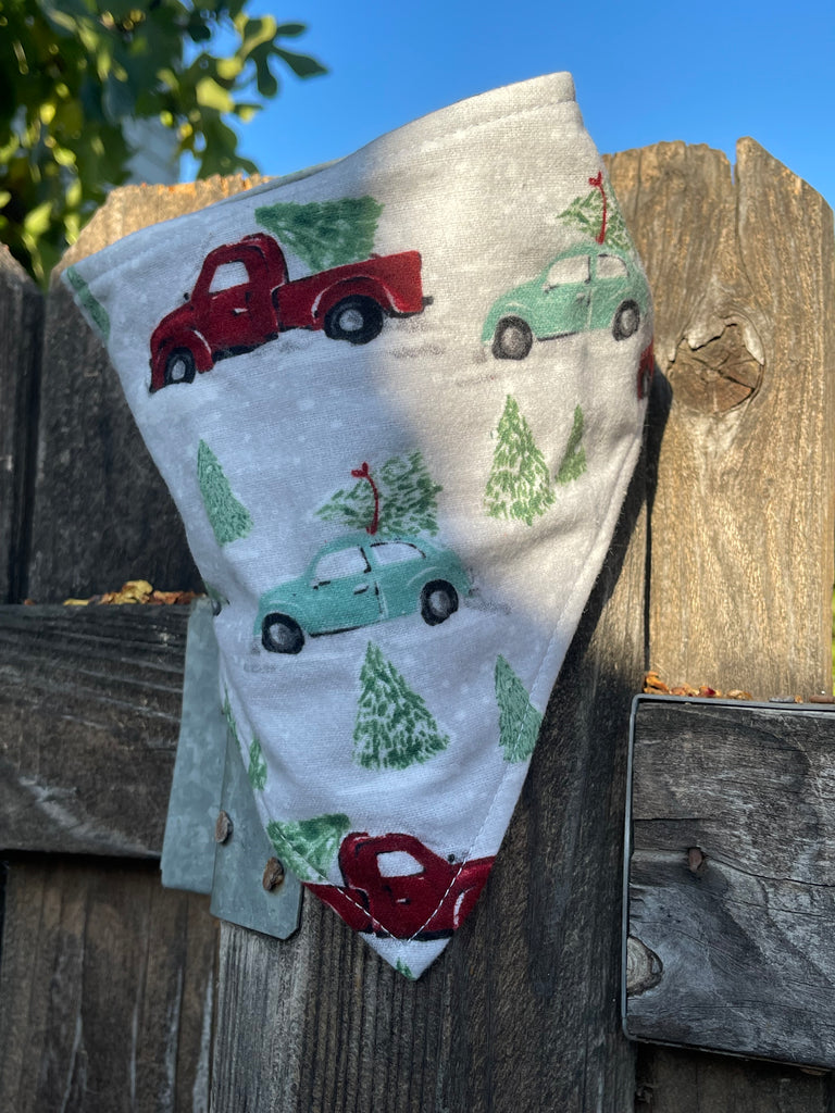 Christmas-themed dog bandana with vintage cars and pine trees resting on a wooden fence in warm sun. Design has a snowy winter scene