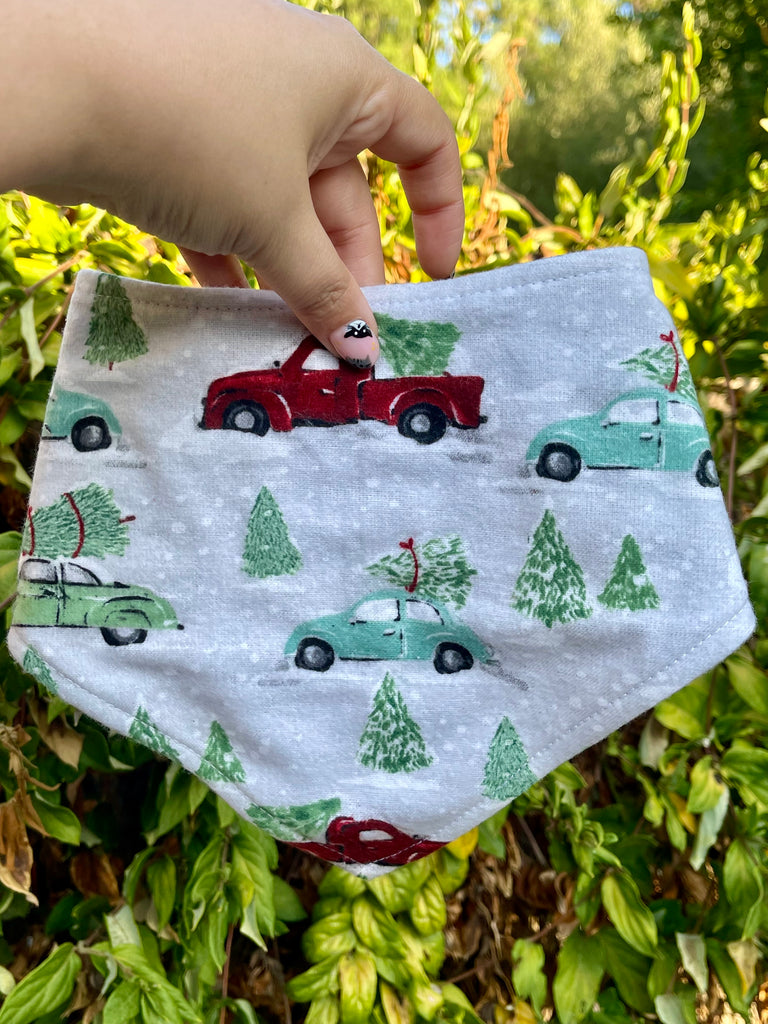 Close-up of the Christmas Tree Farm bandana held in front of green plants. Holiday cars and pine trees are scattered across snowy gray fabric