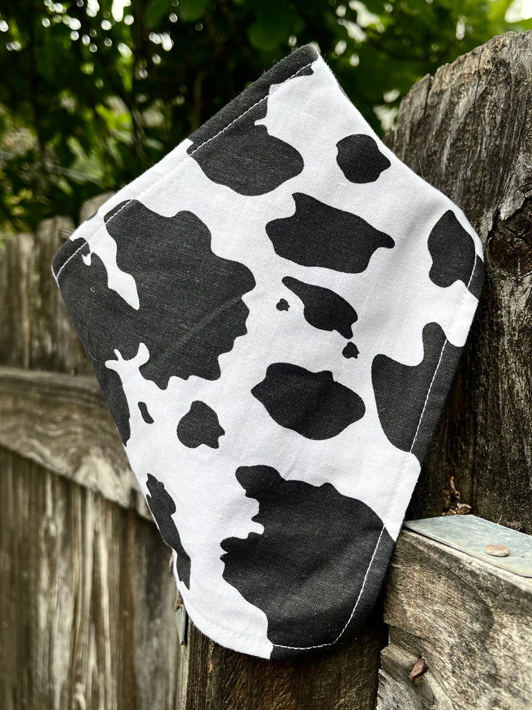 Western-style cow print dog bandana resting on a rustic wooden fence, showcasing bold black and white print and soft handmade texture.