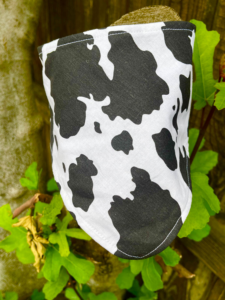 Stylish black and white cowhide bandana displayed upright against tree bark with leafy background, highlighting durable fabric and clean stitching.