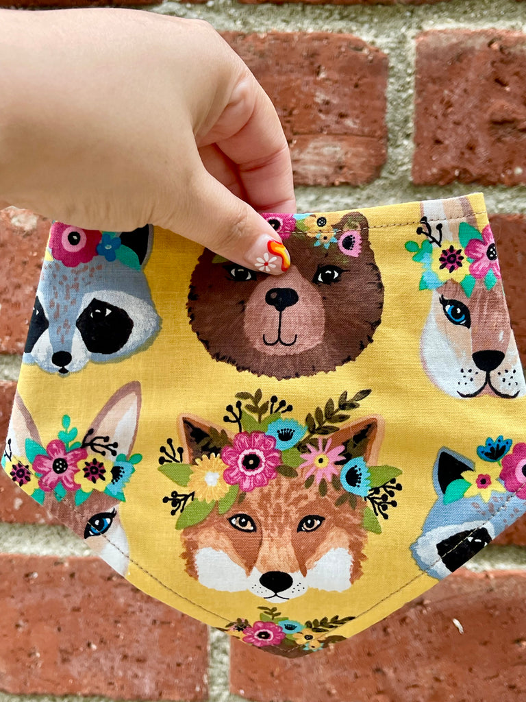 Cute golden pet bandana with whimsical forest animal faces and pink, blue, and green flower crowns, shown in hand against vintage brick wall.