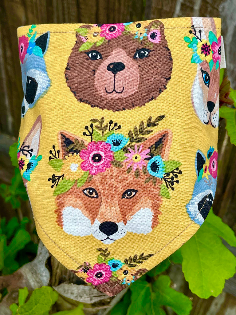 Close-up of eco-friendly yellow pet bandana with woodland critter faces and colorful floral details, displayed on weathered wood fence.