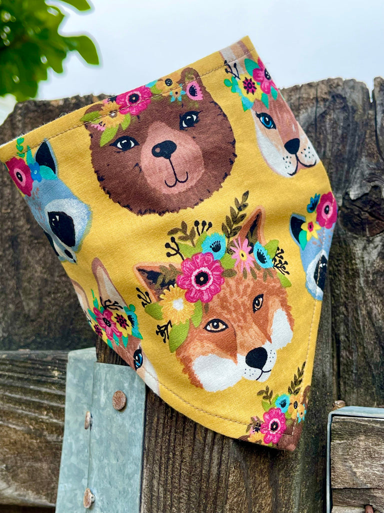 Golden yellow pet bandana featuring fox, bear, raccoon, and floral crown pattern, styled on rustic wooden fence outdoors.