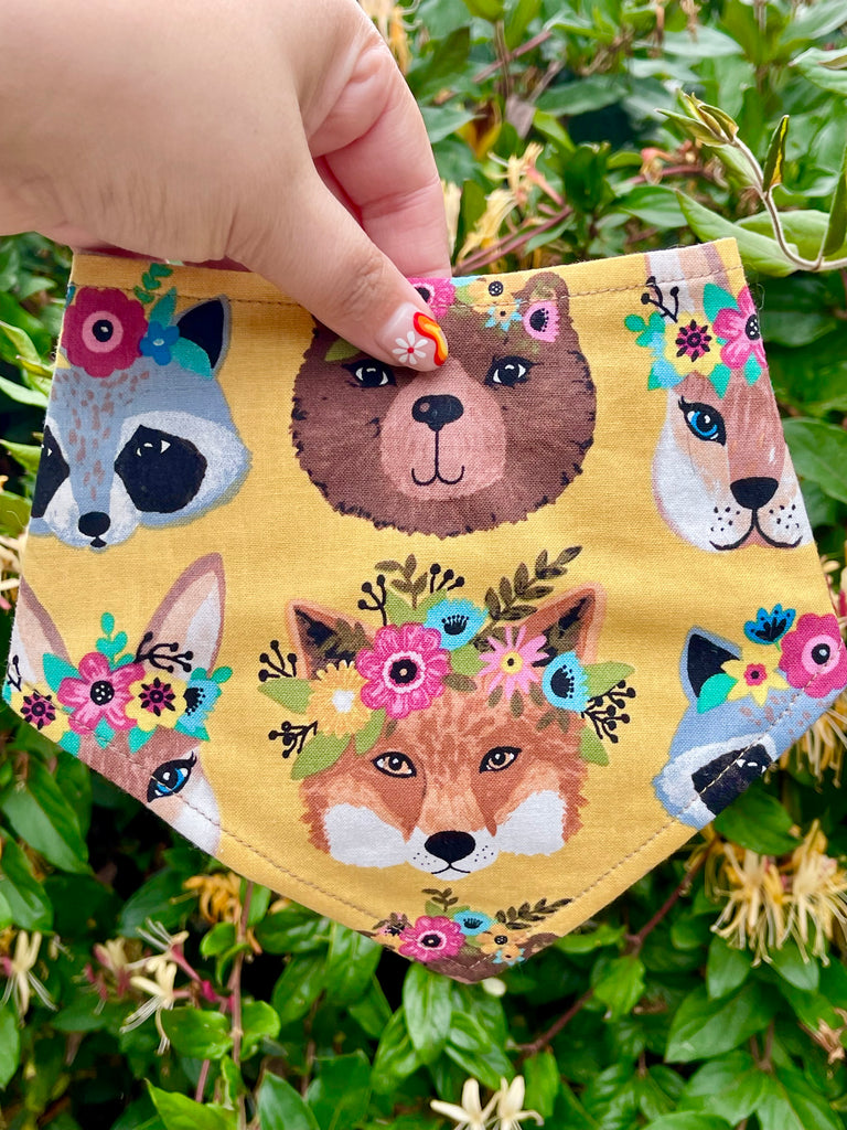 Handmade dog bandana in mustard yellow with bohemian animal faces and bright flower crowns, held by hand with garden background.