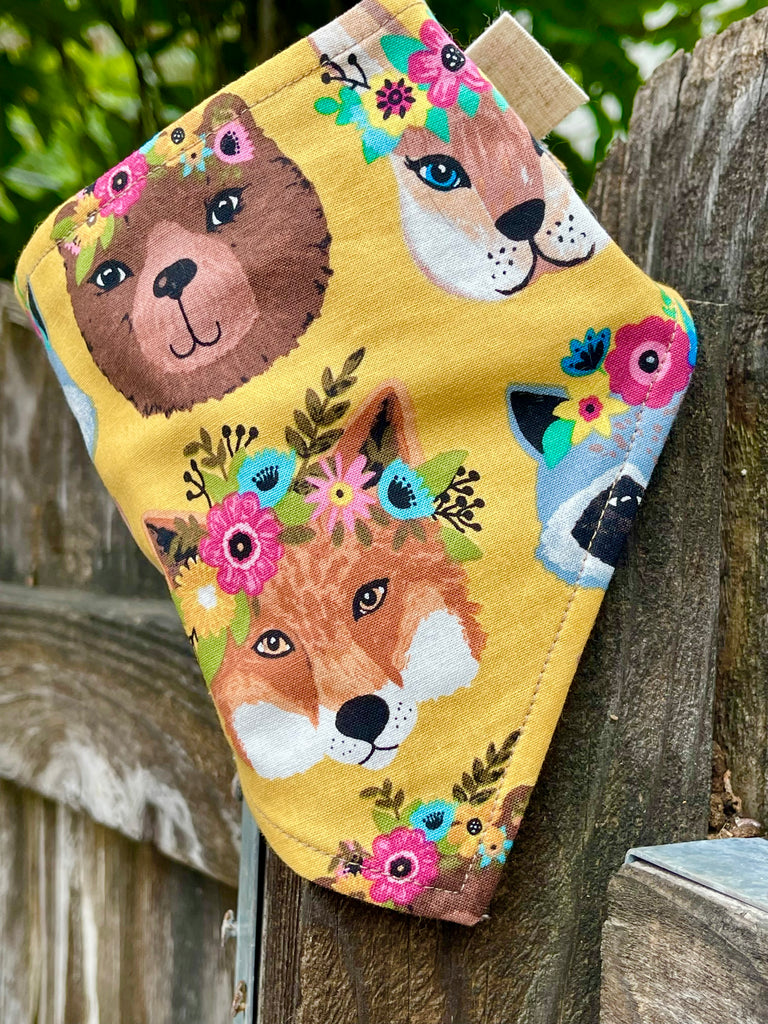 Artisan dog bandana with fox and raccoon floral crown print, golden yellow fabric photographed against natural wood background.