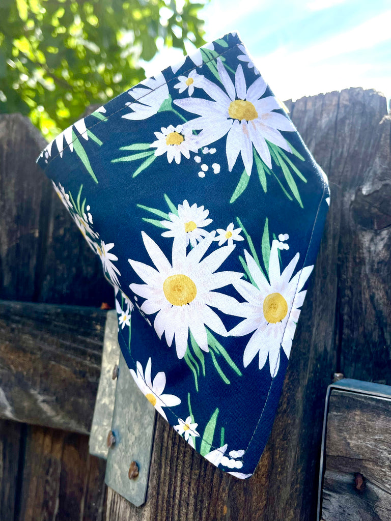 Dog bandana with white and yellow daisy print on navy fabric, hanging on rustic wood fence with blue sky and green leaves in background