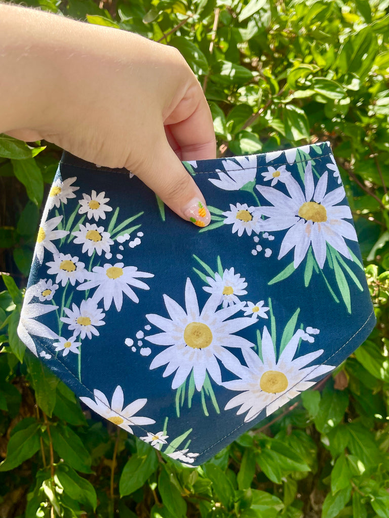 Floral pet bandana with white daisies held in hand in front of leafy green hedge, showing snap closure and bright cheerful design