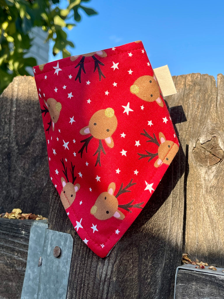 Christmas reindeer bandana draped over a rustic wooden fence in the sun. Features cartoon reindeer and white stars on bold red.
