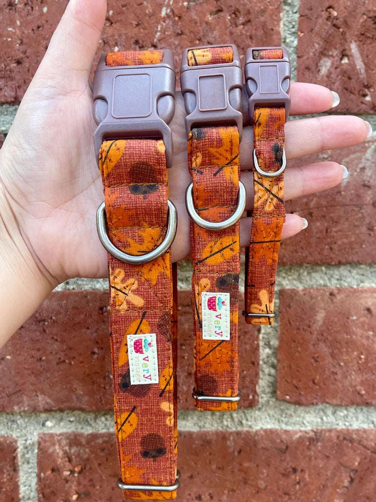 Hand holding three rust-orange plaid dog collars with autumn leaf pattern against vintage brick wall background