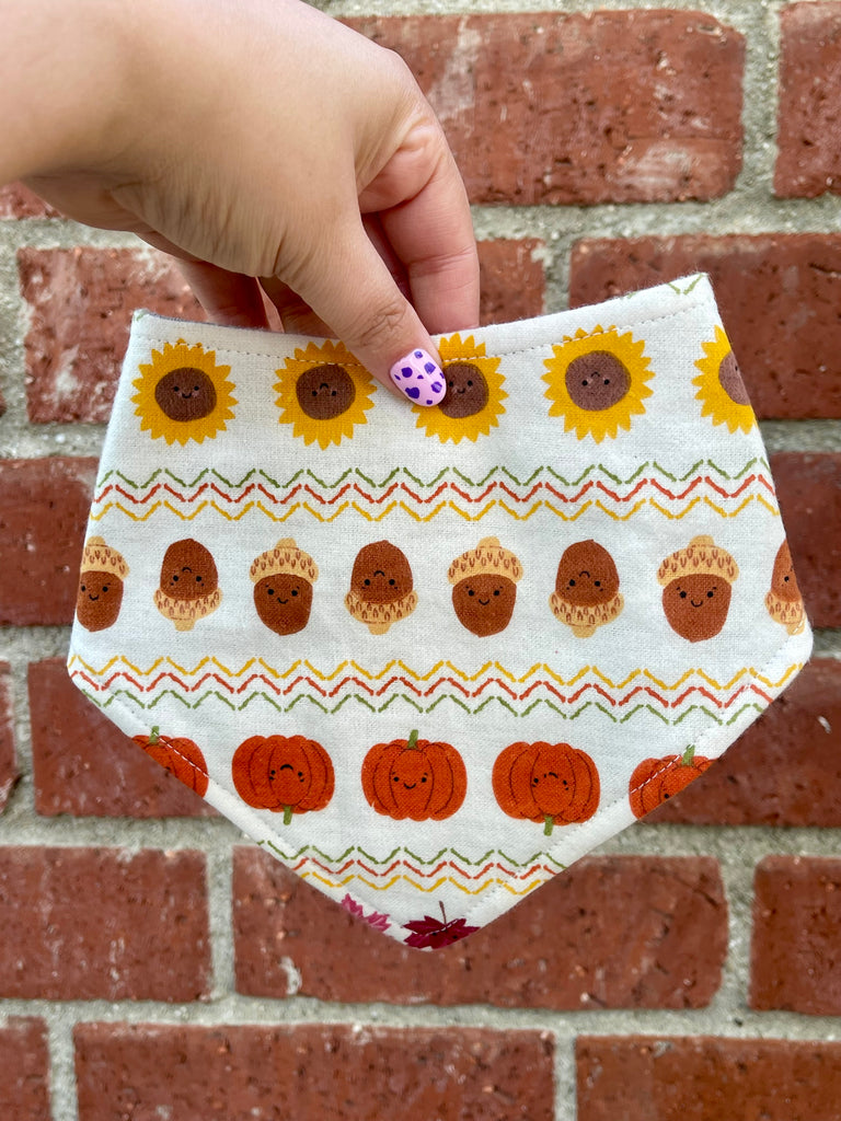 Hand holding a fall dog bandana with rows of smiling sunflowers, acorns, and pumpkins on a cream background in front of a red brick wall.