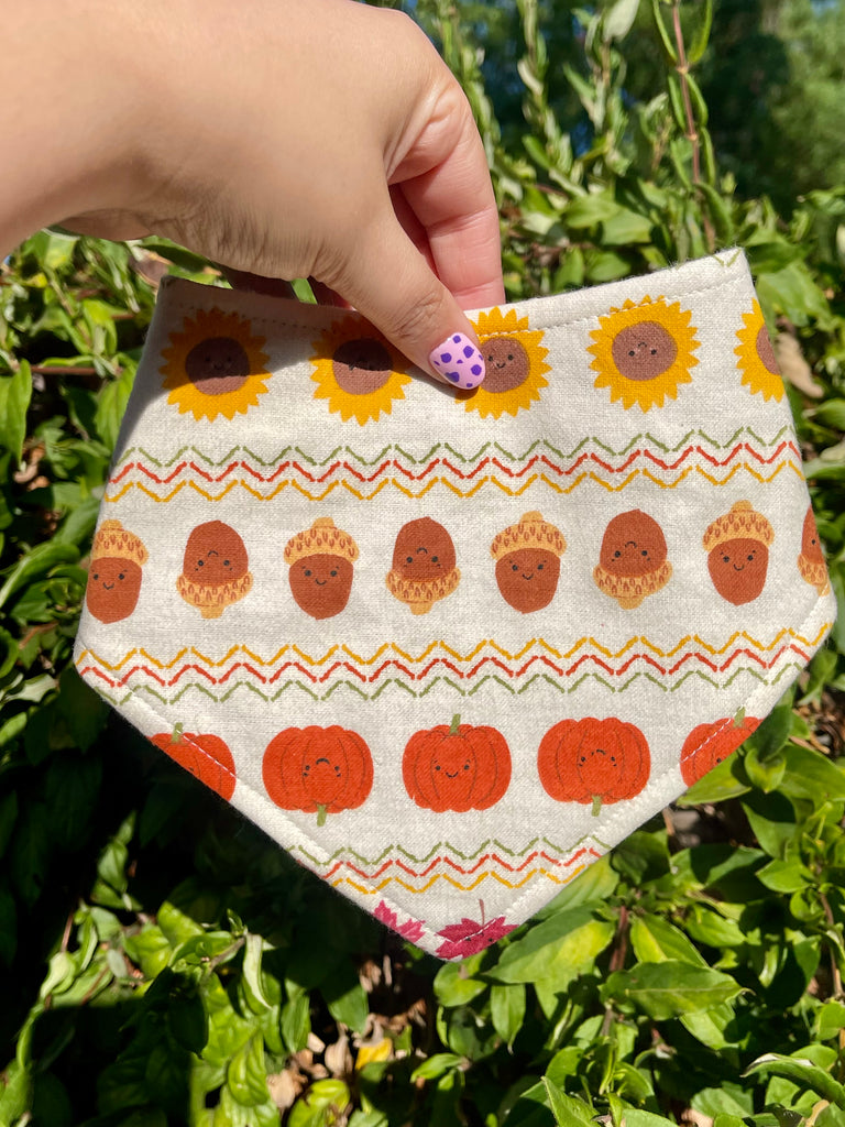 Fall-themed bandana with happy autumn icons including pumpkins, acorns, and sunflowers held up in front of bright green foliage and blue sky.