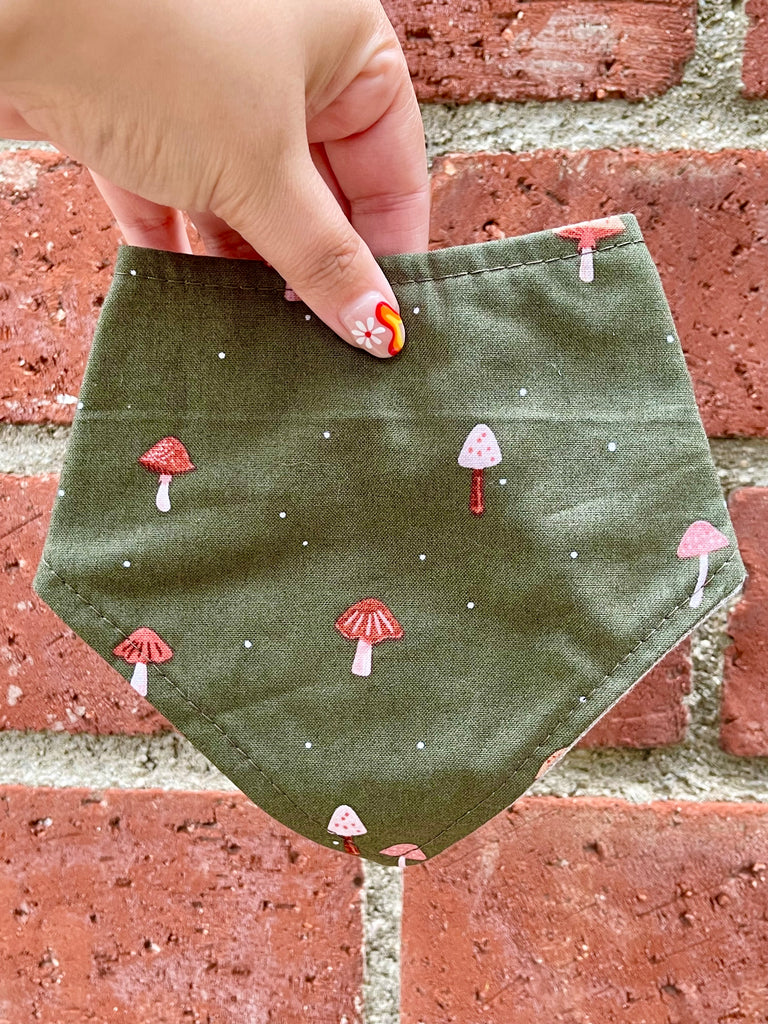 Olive green fall mushroom bandana held in front of red brick wall, showing full pattern and detail. 3 snaps on the back for a secure and adjustable fit.