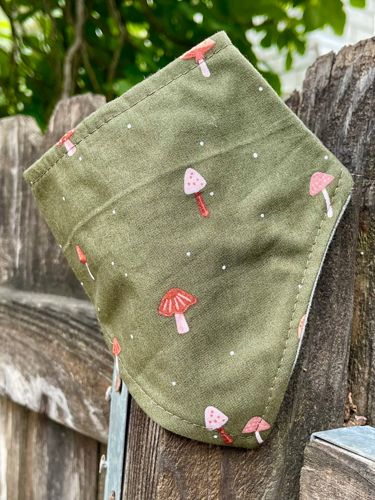 Olive green dog bandana with tiny red and pink mushrooms, shown draped on rustic wood fence with leafy background. Handmade in Los Angeles.
